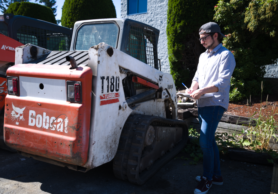 A supervisor in a hard hat and reflective vest carefully diagnoses a skid steer bobcat's issue, exemplifying hands-on expertise and problem-solving in action.